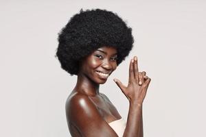 portret van mooi jong Afrikaanse vrouw op zoek Bij camera en gebaren terwijl staand tegen grijs achtergrond foto