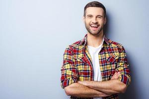 gelukkig jong Mens. portret van knap jong Mens in gewoontjes overhemd houden armen gekruiste en glimlachen terwijl staand tegen grijs achtergrond foto