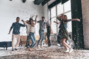 genieten van zorgeloos tijd. vol lengte van gelukkig jong mensen dansen terwijl hebben een partij met confetti vliegend overal foto