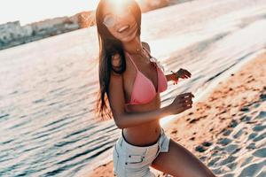 van nature fit en mooi. aantrekkelijk jong vrouw in bikini glimlachen en op zoek weg terwijl wandelen Aan de strand buitenshuis foto