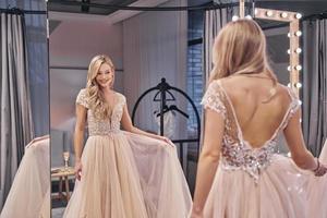 wat een lief jurk. mooi jong vrouw vervelend bruiloft jurk en glimlachen terwijl staand in voorkant van de spiegel in bruids winkel foto