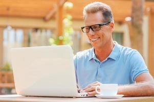 genieten van zijn werk Aan vers lucht. vrolijk volwassen Mens werken Bij laptop en glimlachen terwijl zittend Bij de tafel buitenshuis met huis in de achtergrond foto