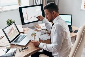 geïnspireerd naar werk moeilijk. jong zakenman in formele kleding vergelijken gegevens gebruik makend van computer terwijl zittend in de kantoor foto