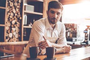 zelfverzekerd barista. jong knap Mens in schort op zoek Bij camera terwijl leunend naar bar teller Bij cafe foto