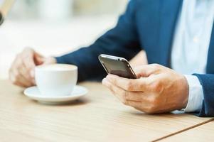 altijd in aanraken. bijgesneden beeld van Mens in formele kleding drinken koffie en typen een bericht Aan mobiel telefoon terwijl zittend in restaurant foto