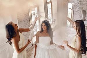 genieten van bruiloft voorbereidende werkzaamheden. top visie van bruidsmeisjes helpen bruid naar zetten Aan een sluier en glimlachen terwijl staand in de passend kamer foto