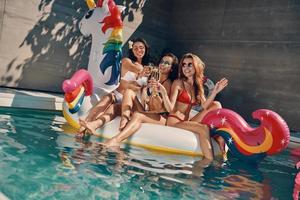 aantrekkelijk jong Dames in badmode glimlachen en drinken Champagne terwijl drijvend Aan opblaasbaar eenhoorn in zwemmen zwembad buitenshuis foto