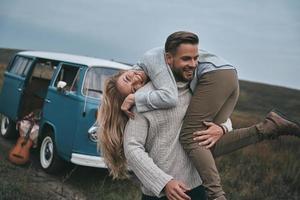 gaan gek. knap jong Mens draag- zijn aantrekkelijk vriendin Aan schouders en glimlachen terwijl staand in de buurt de blauw retro stijl mini busje foto