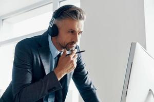 attent volwassen zakenman in hoofdtelefoons op zoek Bij de computer terwijl staand in kantoor foto