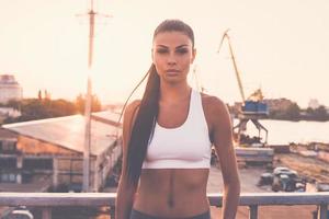 klaar voor opleiding. mooi jong vrouw in sport- kleding op zoek Bij camera terwijl staand Aan de brug met stedelijk visie in de achtergrond foto