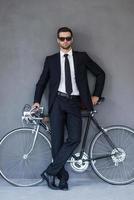 zakenman met zijn fiets. vol lengte van knap jong zakenman leunend Bij de fiets en op zoek Bij camera terwijl staand tegen grijs achtergrond foto