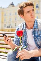 aan het wachten voor haar. bezorgd jong Mens Holding single roos en mobiel telefoon terwijl zittend Aan de bank foto