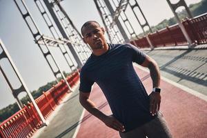 sterk atleet. knap jong Afrikaanse Mens in sport- kleding op zoek Bij camera terwijl staand Aan de brug buitenshuis foto