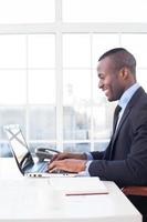 zakenman Bij werk. kant visie van vrolijk jong Afrikaanse zakenman gebruik makend van computer en glimlachen terwijl zittend Bij zijn werken plaats foto