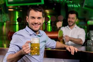 Mens in bier kroeg. knap jong Mens Holding een bier mok en glimlachen terwijl barman gieten bier Aan de achtergrond foto