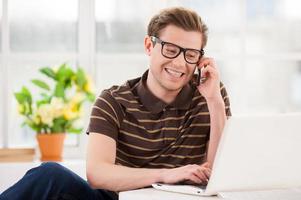 werken Bij huis. knap jong Mens in bril werken Aan laptop en pratend Aan de mobiel telefoon terwijl zittend Aan de verdieping Bij zijn appartement foto