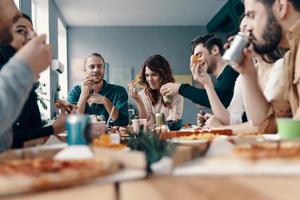 genieten van pizza met vrienden. groep van jong mensen in gewoontjes slijtage aan het eten pizza en glimlachen terwijl hebben een avondeten partij binnenshuis foto