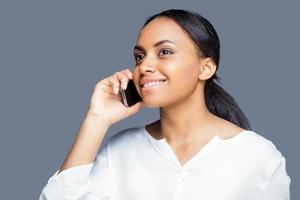schoonheid Aan de telefoon. mooi jong Afrikaanse vrouw pratend Aan de mobiel telefoon en glimlachen terwijl staand tegen grijs achtergrond foto