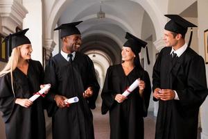 pratend over helder toekomst. vier college afgestudeerden in diploma uitreiking jurken wandelen langs Universiteit gang en pratend foto