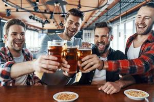 gelukkig jong mannen in gewoontjes kleding roosteren elk andere met bier en lachend terwijl zittend in de kroeg foto