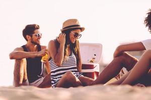 strand tijd met vrienden. vrolijk jong mensen uitgeven mooi hoor tijd samen terwijl zittend Aan de strand en drinken bier foto