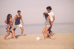 strand pret met vrienden. groep van vrolijk jong mensen spelen met voetbal bal Aan de strand met zee in de achtergrond foto