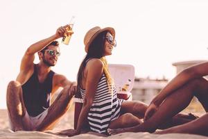 uitgeven tijd met vrienden. vrolijk jong mensen uitgeven mooi hoor tijd samen terwijl zittend Aan de strand en drinken bier foto