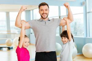 trots naar worden sterk. vrolijk jong sportief vader tonen zijn biceps terwijl kinderen bonding naar hem en glimlachen foto
