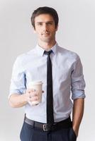 koffie pauze. knap jong Mens in overhemd en stropdas Holding koffie kop en op zoek Bij camera terwijl staand tegen grijs achtergrond foto