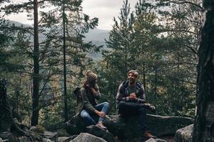 Super goed reis. mooi jong paar zittend Aan de rotsen en glimlachen terwijl wandelen samen in de bossen foto