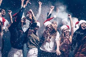 Kerstmis plezier. groep van mooi jong mensen in de kerstman hoeden het werpen kleurrijk confetti en op zoek gelukkig foto