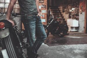 Mens in de buurt zijn fiets. zelfverzekerd jong Mens houden armen gekruiste en op zoek Bij camera terwijl staand in de buurt zijn motorfiets garage of reparatie winkel foto