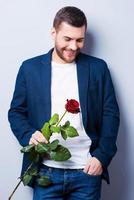 elegant romantisch. knap jong Mens Holding een bloem terwijl staand tegen grijs achtergrond foto