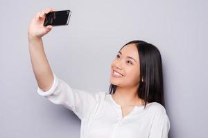 selfie vrolijk jong Aziatisch vrouw Holding mobiel telefoon en maken selfie door haar slim telefoon terwijl staand tegen grijs achtergrond foto