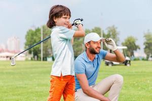 mooi hoor schot zelfverzekerd weinig jongen spelen golf terwijl zijn vader staand dichtbij naar hem Aan de golf Cursus foto