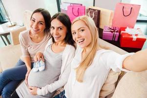 selfie top visie van gelukkig jong zwanger vrouw Holding baby slofjes Aan haar buik en glimlachen terwijl twee vrienden zittend dichtbij naar haar en maken selfie foto