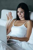 mooi hoor naar zien u online jong vrouw glimlachen en golvend terwijl op zoek Bij haar laptop en zittend in de bed Bij huis foto