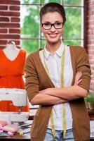 zelfverzekerd kleermaker. mooi jong vrouw kleermaker Holding schaar en glimlachen Bij camera terwijl mannequin staand in de achtergrond foto