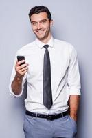 zakenman met mobiel telefoon. zelfverzekerd jong Mens in overhemd en stropdas Holding mobiel telefoon en glimlachen terwijl staand tegen grijs achtergrond foto