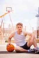 basketbal speler. zelfverzekerd jong mannetje basketbal speler zittend buitenshuis met bal foto