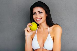 eten gezond aantrekkelijk jong vrouw in wit bikini Holding groen appel en glimlachen terwijl staand tegen grijs achtergrond foto