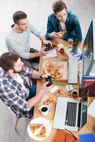 ontspannende na werk. top visie van drie jong mannen spelen computer spellen en aan het eten pizza terwijl zittend Bij de bureau foto