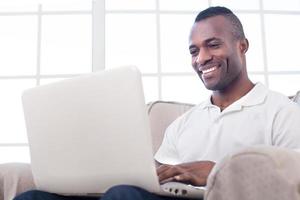 werken Bij huis. vrolijk Afrikaanse Mens gebruik makend van computer en glimlachen terwijl zittend Aan de stoel foto