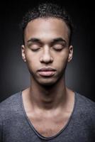 kalmte en zelfverzekerd. portret van jong Afrikaanse Mens houden ogen Gesloten terwijl staand tegen zwart achtergrond foto
