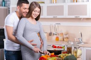 Koken gezond voedsel. gelukkig jong paar Koken samen in de keuken terwijl Mens aanraken buik van zijn zwanger vrouw foto