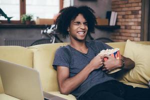 lol. vrolijk jong Afrikaanse Mens aan het kijken TV en Holding emmer van popcorn terwijl zittend Aan de bankstel Bij huis foto