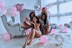 tonen glimlach vier aantrekkelijk jong Dames in pyjama glimlachen terwijl nemen selfie in de slaapkamer met ballonnen allemaal over- de plaats foto