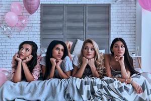 proberen naar vind een antwoord verward vier jong Dames in pyjama op zoek weg terwijl aan het liegen Aan de bed foto