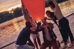 voorbereidingen treffen papier lantaarn. gelukkig jong mensen in gewoontjes slijtage verlichting omhoog lucht lantaarn terwijl staand Aan de pier foto