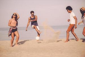 zomer pret met vrienden. groep van vrolijk jong mensen spelen met voetbal bal Aan de strand met zee in de achtergrond foto